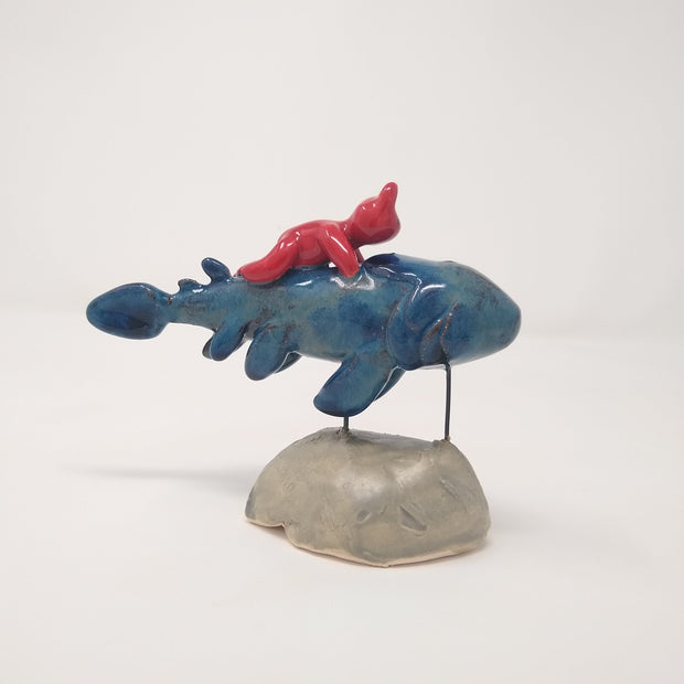 Ceramic sculpture of a blue fish with many fins and no facial features. A small red devil, also without any facial features, rides atop of it. The pair are held up by 2 pieces of wire that lead to a gray mound that resembles stone.