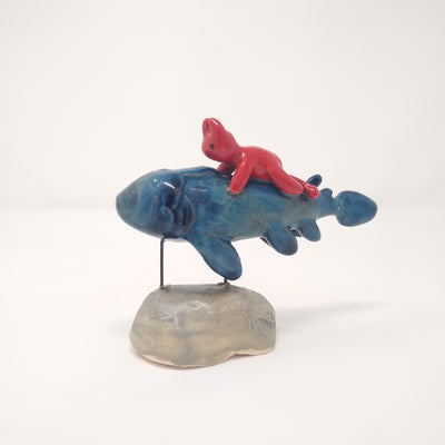 Ceramic sculpture of a blue fish with many fins and no facial features. A small red devil, also without any facial features, rides atop of it. The pair are held up by 2 pieces of wire that lead to a gray mound that resembles stone.