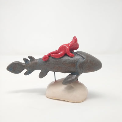 Ceramic sculpture of a grey fish with many fins and no facial features. A small red devil, with minimal facial features, rides atop of it. The pair are held up by 2 pieces of wire that lead to a cream colored mound that resembles stone.