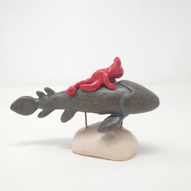 Ceramic sculpture of a grey fish with many fins and no facial features. A small red devil, with minimal facial features, rides atop of it. The pair are held up by 2 pieces of wire that lead to a cream colored mound that resembles stone.