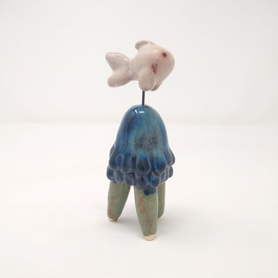 Ceramic sculpture of a blue jellyfish like creature, with 3 legs like a table. Mounted above it via a wire is a white fish, akin to a goldfish.