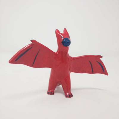 Shiny red ceramic sculpture of Rodan, a dragon like monster with a dark blue beak and extended wings. It stands on 2 legs.