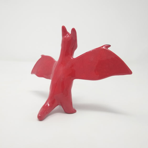 Shiny red ceramic sculpture of Rodan, a dragon like monster with a dark blue beak and extended wings. It stands on 2 legs. Back view.