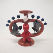 Ceramic sculpture of a small red devil on sitting on a brown vessel, with small blue balls coming out of it and 2 black bats, facing the devil. Over its head is a small white ghost. 