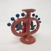 Ceramic sculpture of a small red devil on sitting on a brown vessel, with small blue balls coming out of it and 2 black bats, facing the devil. Over its head is a small white ghost. Back view.