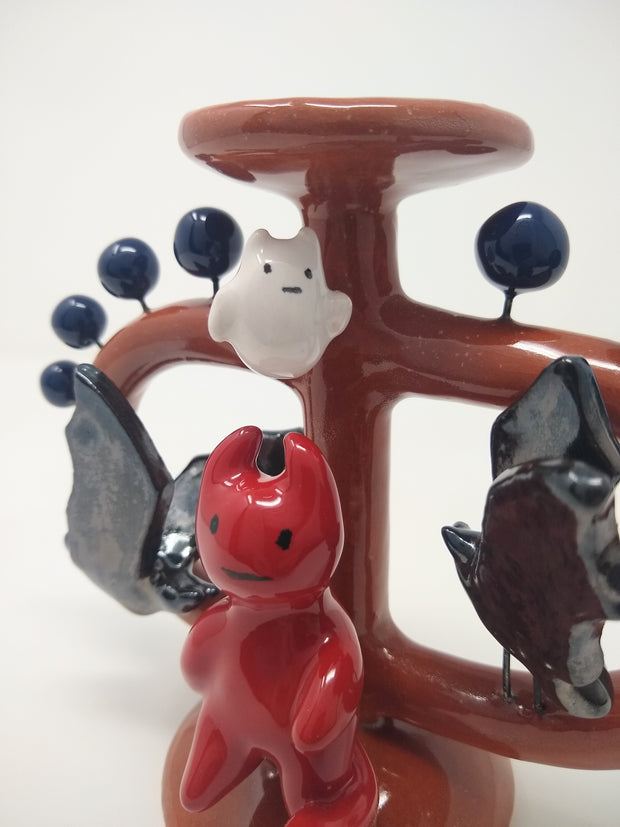Ceramic sculpture of a small red devil on sitting on a brown vessel, with small blue balls coming out of it and 2 black bats, facing the devil. Over its head is a small white ghost. Close up.