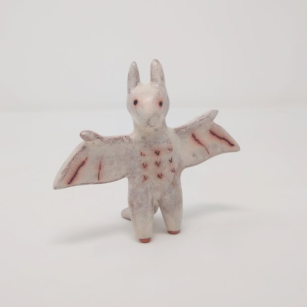 Ceramic sculpture of Rodan, a dragon like monster with tall ears like a bat and extended wings. It is an off white color with simplistic drawn on details, the color and effect of rust. It stands on 2 legs.