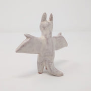 Ceramic sculpture of Rodan, a dragon like monster with tall ears like a bat and extended wings. It is an off white color with simplistic drawn on details, the color and effect of rust. It stands on 2 legs. Back view.