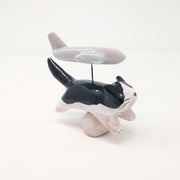 Ceramic sculpture of a tuxedo colored cat, mid leap. It is positioned over a small white mound and coming out of its back is a simplistic plane, attached by wire.
