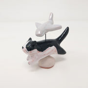 Ceramic sculpture of a tuxedo colored cat, mid leap. It is positioned over a small white mound and coming out of its back is a simplistic plane, attached by wire. 