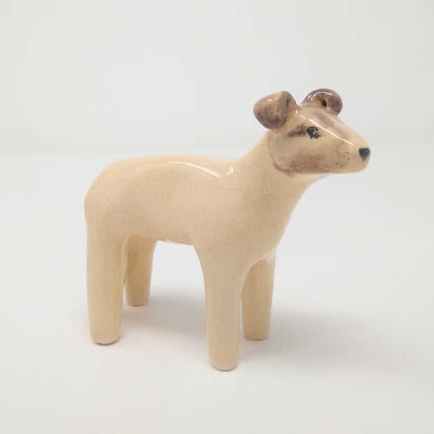 Glazed ceramic sculpture of a cream colored dog with a tan face, it has simplistic limbs and drawn on facial features.