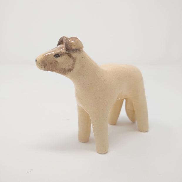 Glazed ceramic sculpture of a cream colored dog with a tan face, it has simplistic limbs and drawn on facial features.