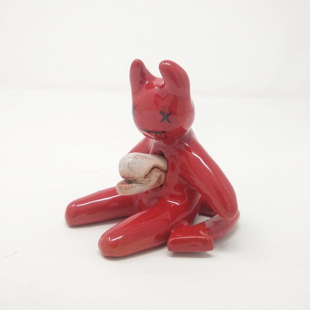 Shiny, glazed red ceramic sculpture of a red devil sitting on the ground, with a white alien bursting from its chest. The devil's face has x's for eyes and a tongue out expression.