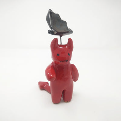 Glazed red sculpture of a small devil, with simplistic body parts and a long pointed tail. Atop its head, attached with wire is a black bat, also with simplistic body shapes.