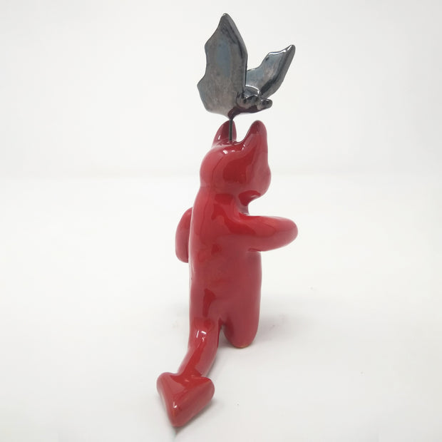 Glazed red sculpture of a small devil, with simplistic body parts and a long pointed tail. Atop its head, attached with wire is a black bat, also with simplistic body shapes.