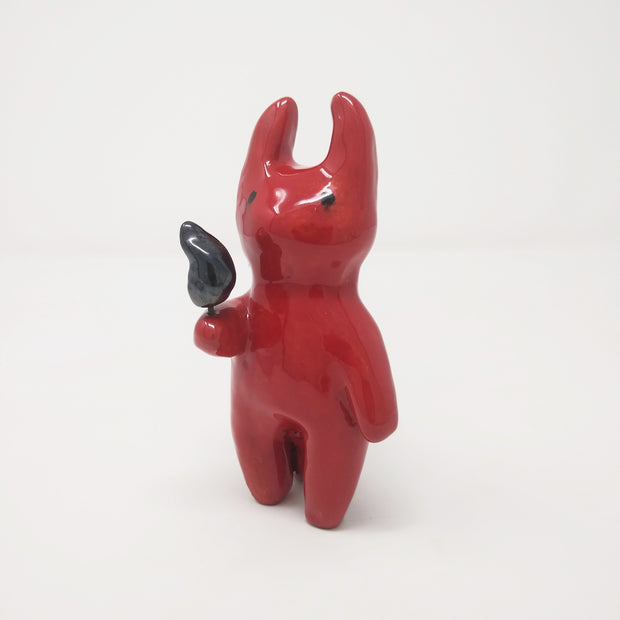 Glazed red ceramic sculpture of a devil with simplistic body shapes and simple black teary eyes. It holds a small black flame in one hand.