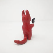Glazed red ceramic sculpture of a devil with simplistic body shapes and simple black teary eyes. It holds a small black flame in one hand.