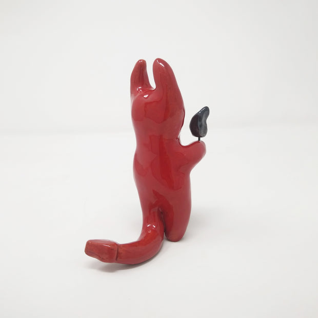 Glazed red ceramic sculpture of a devil with simplistic body shapes and simple black teary eyes. It holds a small black flame in one hand.