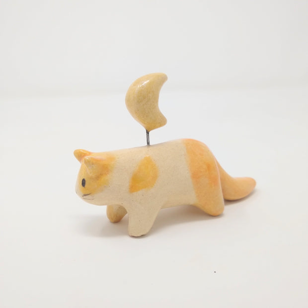 Ceramic sculpture of a small orange spotted cat, with simplistic limbs and drawn on eyes and whiskers. Attached to its back, via a wire, is a crescent moon.