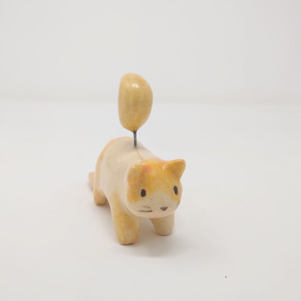 Ceramic sculpture of a small orange spotted cat, with simplistic limbs and drawn on eyes and whiskers. Attached to its back, via a wire, is a crescent moon.