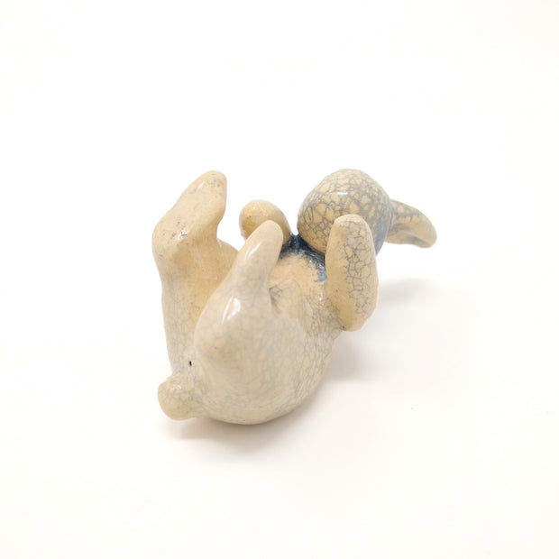 Off white ceramic sculpture with a crackled outer glaze of a rabbit, laying on its back with its feet up in the air and arms reaching its toes. Subtle blue coloring surrounds its neck and face.