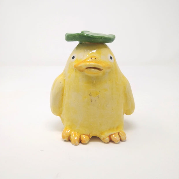 Ceramic sculpture of a yellow duck with a green leaf atop its head. 