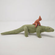 Unglazed ceramic sculpture of a long komodo dragon, with a very long tail. Atop its back is a small red devil like character, riding on top.