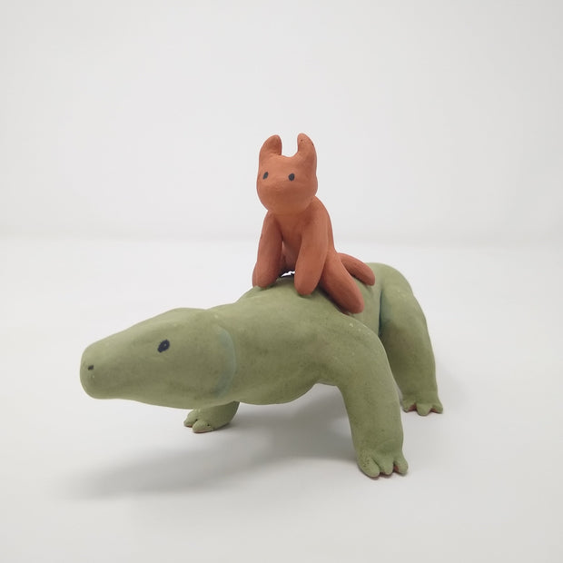 Unglazed ceramic sculpture of a long komodo dragon, with a very long tail. Atop its back is a small red devil like character, riding on top.