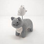 Ceramic sculpture of a gray cat with simplistic body features and a drawn on eyes and whiskers. Hovering atop its back is a small white ghost, with watercolor style tears.