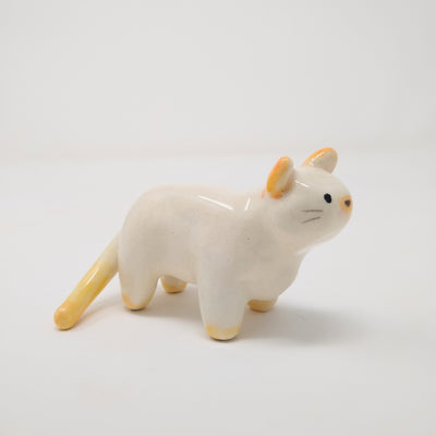 White ceramic cat with simplistic bodily features and a drawn on eyes and nose. Atop its ears, tail and feet is a subtle orange coloring.