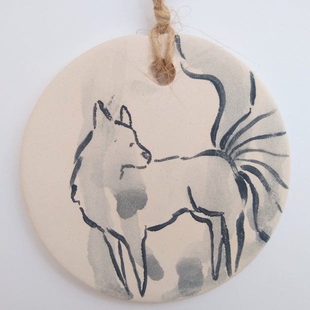 Painting on a circular flat piece of glazed ceramic of a line art fox with flowing tails.