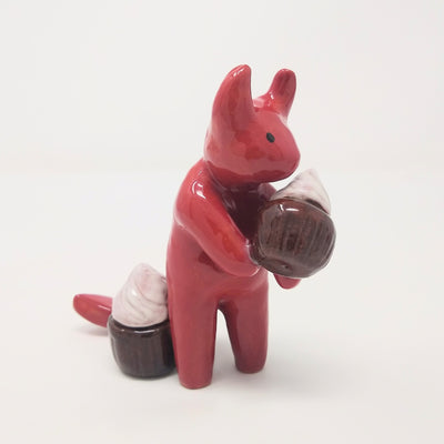 Ceramic sculpture of a red devil character with simplified body features, standing and holding a chocolate cupcake. Another cupcake sits near its tail.