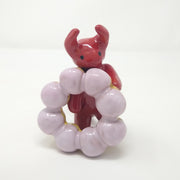Sculpture of a small red devil character with simplistic body shapes and only eyes for facial features. It holds a large mochi donut with light purple frosting.