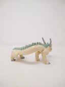 Ceramic sculpture of a white dragon standing on short legs with a very long tail and mint green short spikes along its back.