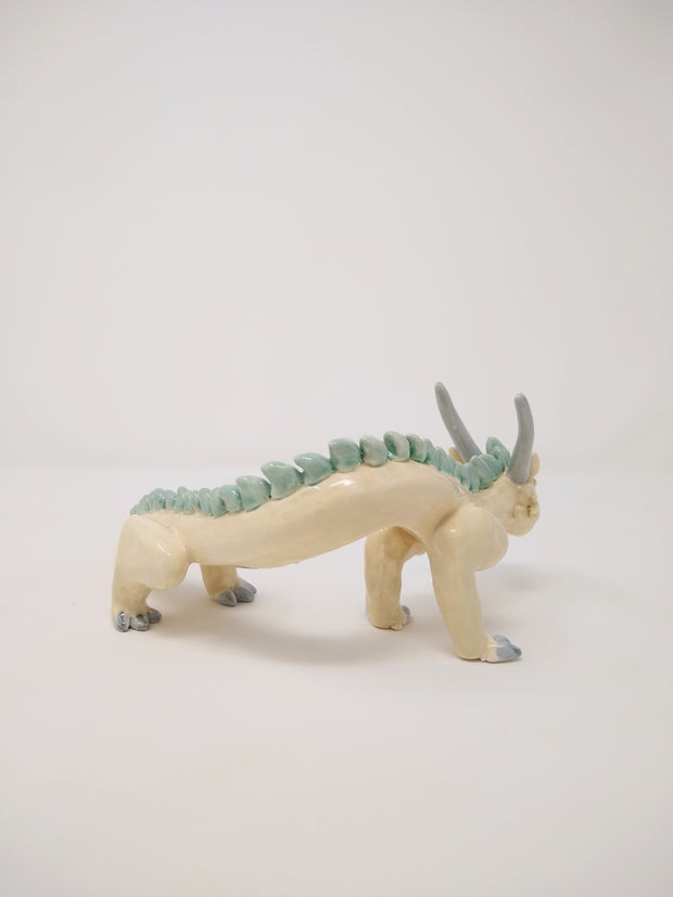 Ceramic sculpture of a white dragon standing on short legs with a very long tail and mint green short spikes along its back.