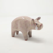 Ceramic sculpture of a light pinkish tan colored boar, with drawn on eyes and white tusks. It stands with a slightly hunched back.