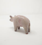 Ceramic sculpture of a light pinkish tan colored boar, with drawn on eyes and white tusks. It stands with a slightly hunched back.