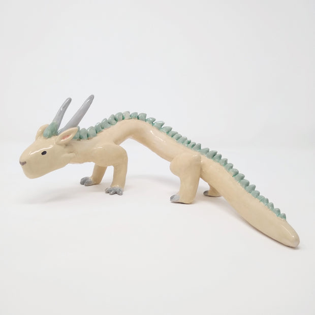 Ceramic sculpture of a white dragon standing on short legs with a very long tail and mint green short spikes along its back.