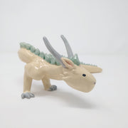 Ceramic sculpture of a white dragon standing on short legs with a very long tail and mint green short spikes along its back.