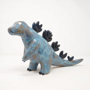 Ceramic sculpture of a brown and blue abstract Godzilla figure, with abstract dark blue spikes on its back, no facial features, and one arm extended out in front.