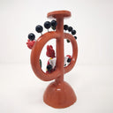 Ceramic sculpture of a small red devil sitting on a brown vessel with 2 large handles and a skinny middle. Small leaves and flowers decorate the vessel with black circles protruding out via wire.