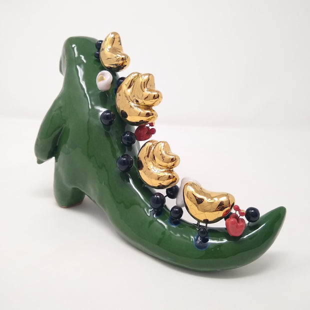 Sculpture of a green Godzilla like monster, standing with its arms slightly out in front of it. Its back and tail are lined with various abstract sculptural shapes and small balls, consisting of gold, black and red and white flowers.