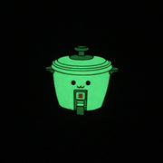 Die cut enamel pin of a rice cooker, with a small simple set of eyes and a cute smile. Pin is glowing in the dark.