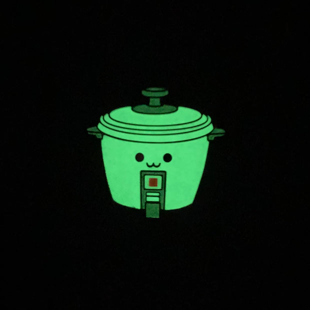 Die cut enamel pin of a rice cooker, with a small simple set of eyes and a cute smile. Pin is glowing in the dark.