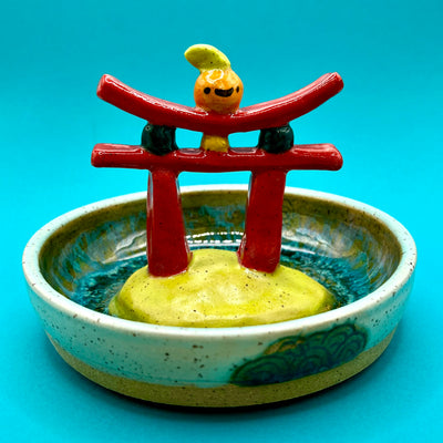 Ceramic sculpture of a red gate with a small orange citrus on top, within a ceramic bowl.