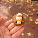 Hand holding a small figurine of a dog with a white body and brown face on a wooden base, with a blurred outdoor background.
