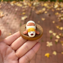 Hand holding a small figurine of a dog with a white body and brown face on a wooden base, with a blurred outdoor background.