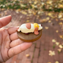Hand holding a small figurine of a hot dog with mustard on a wooden base against a blurred outdoor background.