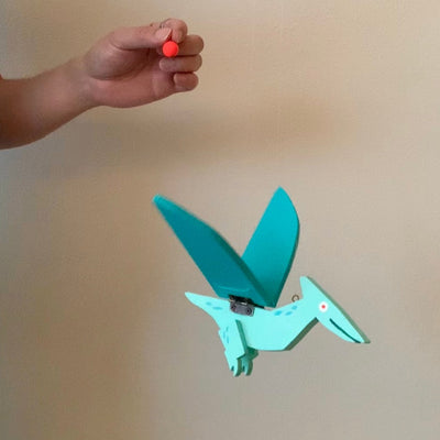 Teal colored wooden puppet of a pterodactyl with a cartoon expression: a simple smile and a white eye with a red dot. Its wings flap up and down, pulled by a string.