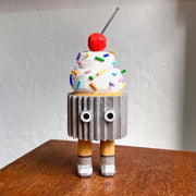 Wooden sculpture of a cupcake with white frosting, sprinkles, and a cherry on top. Its wrapped in a silver wrapper and stands on 2 legs, with a pair of eyes as its only facial feature.